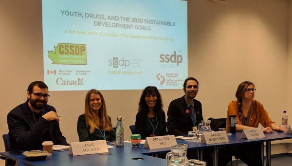 group of five people seated at a table, with logos of drug policy organizations behind them. Name tags read: Jake Agliata, Penelope Hill, Nazlee Maghsoudi, Alex Betsos (the nametag of the fifth person, Orsi Fehér, is out of frame)